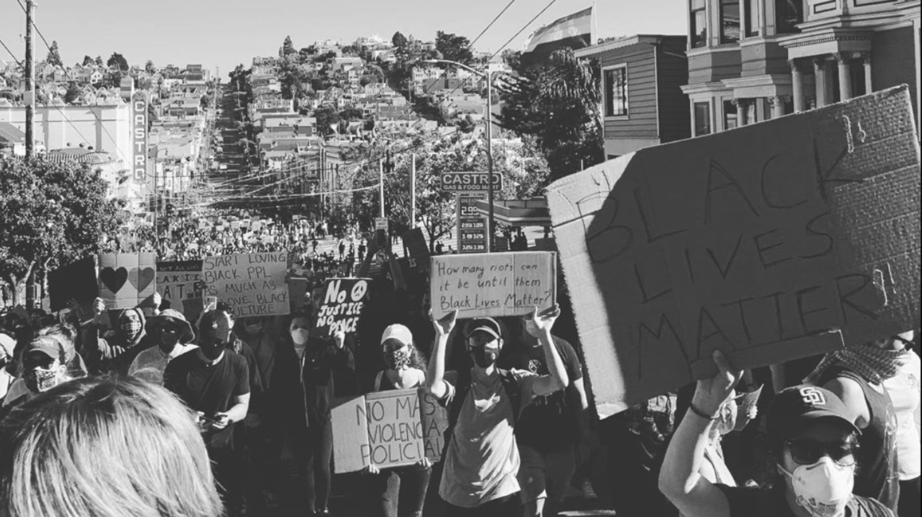A photo of people marching down a street holding signs including No Justice No Peace at a Black Lives Matter protest in San Francisco organized by 17 year-old Simone Jacques