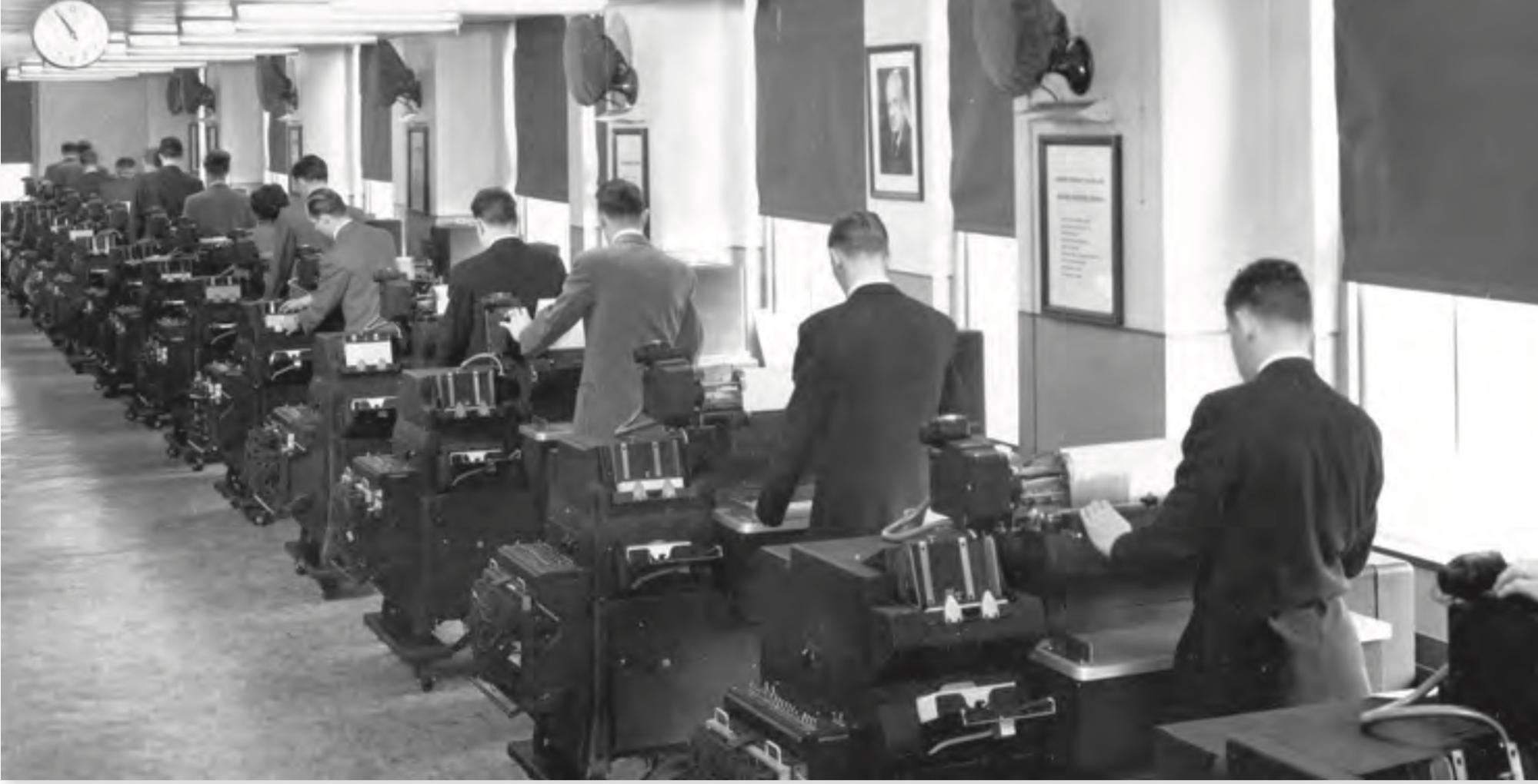 Black-and-white photograph depicting a row of suit-wearing men standing in front of their desks, operating old computer equipment.
