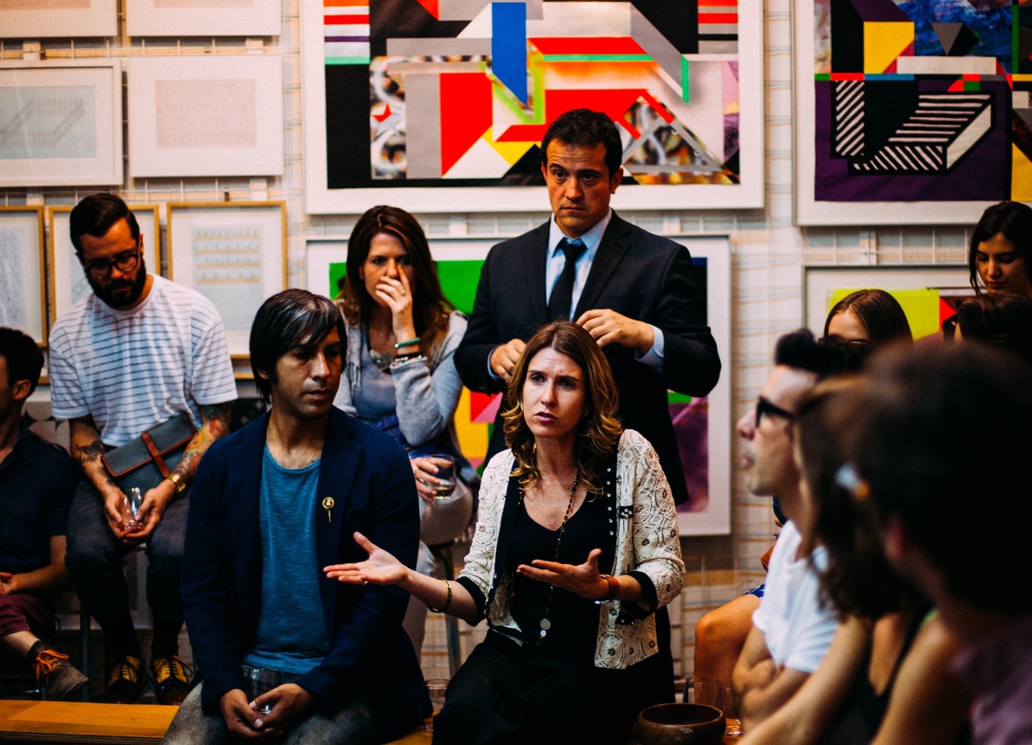 A person with long hair and gesturing with hands out appears to share a story in a room with a dozen other people in business casual attire and modern art on the wall behind them