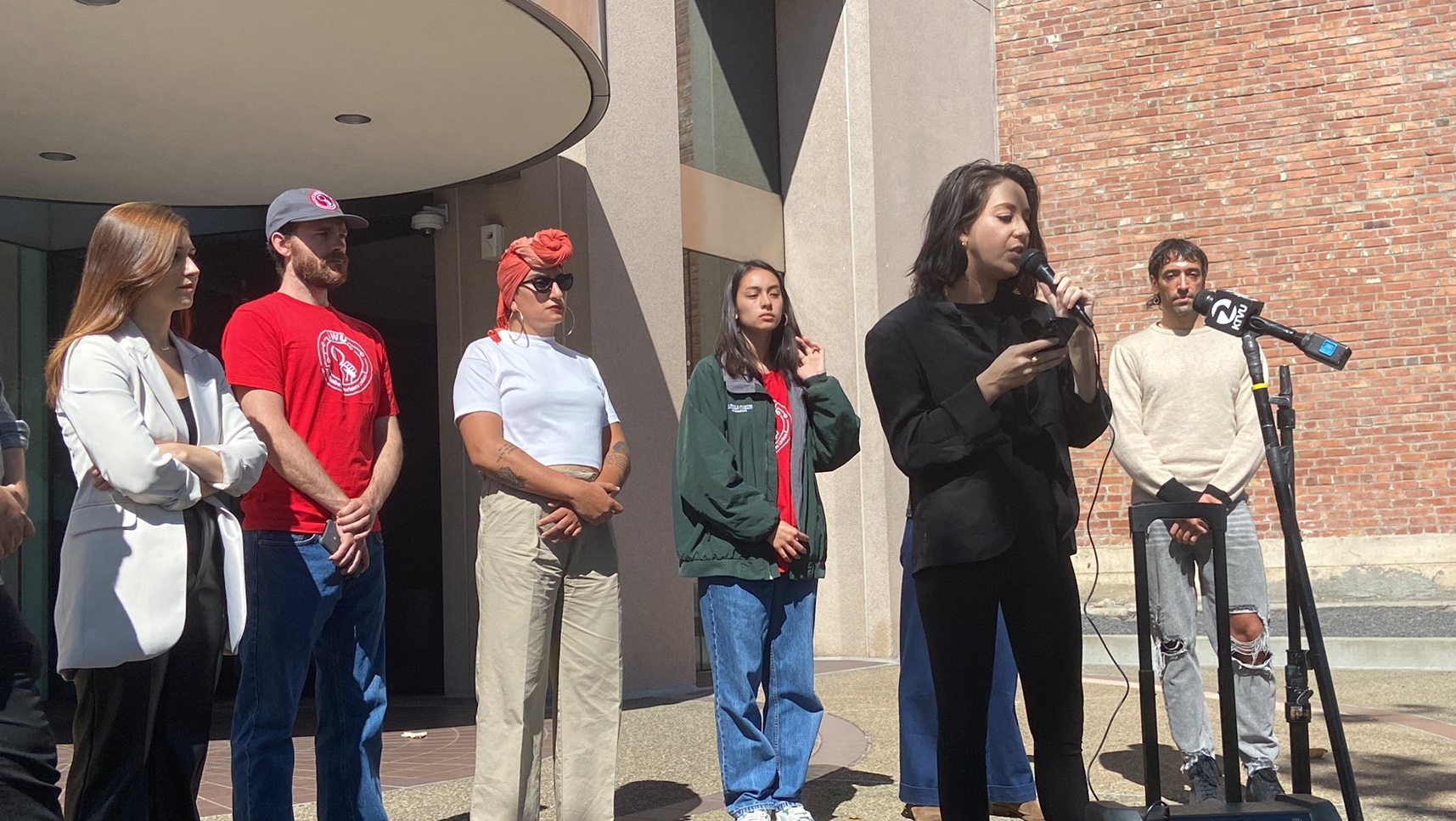 Eight Alphabet workers in casual clothing speaking on a mic in front of headquarters with a brick wall behind them