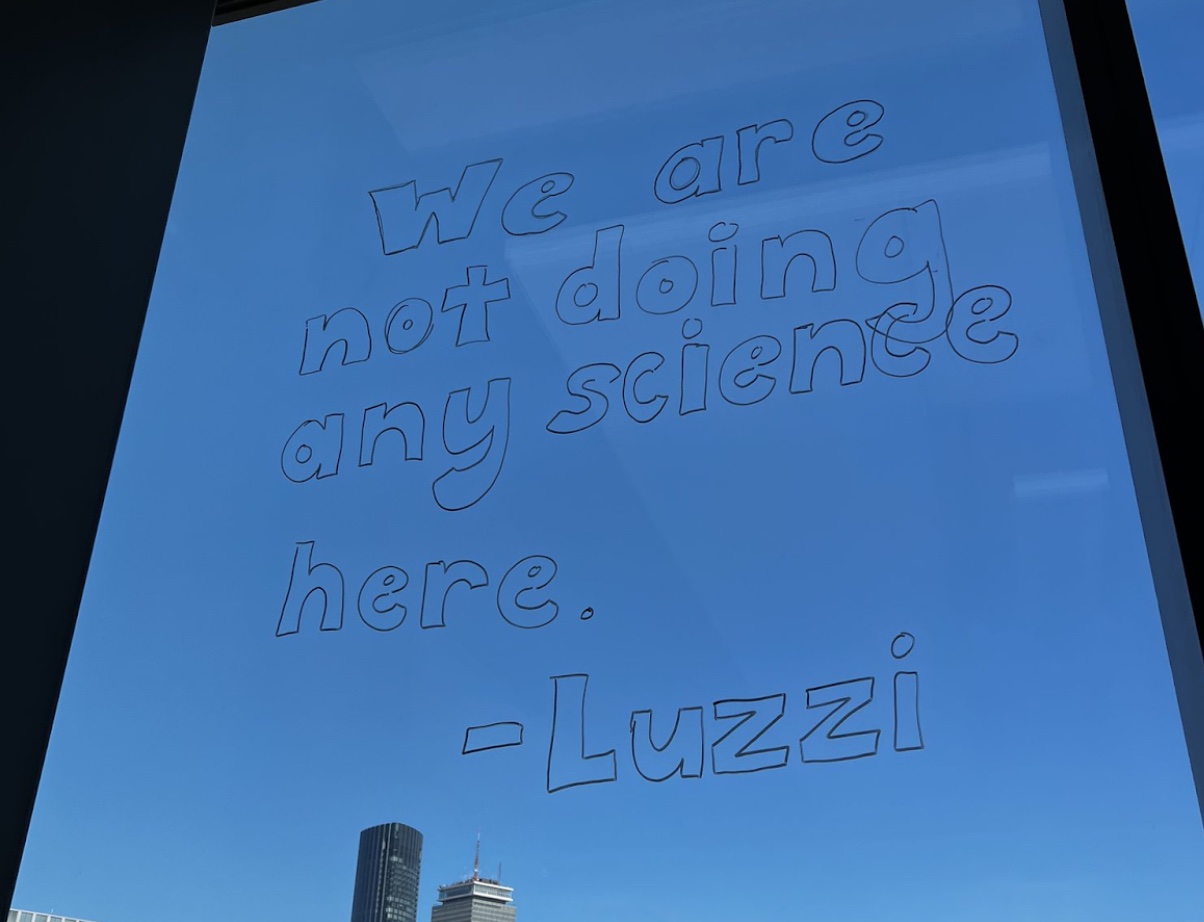 A window looking out onto the Boston skyline and a clear blue sky with writing in dry-erase marker that reads, WE ARE NOT DOING ANY SCIENCE HERE - LUZZI