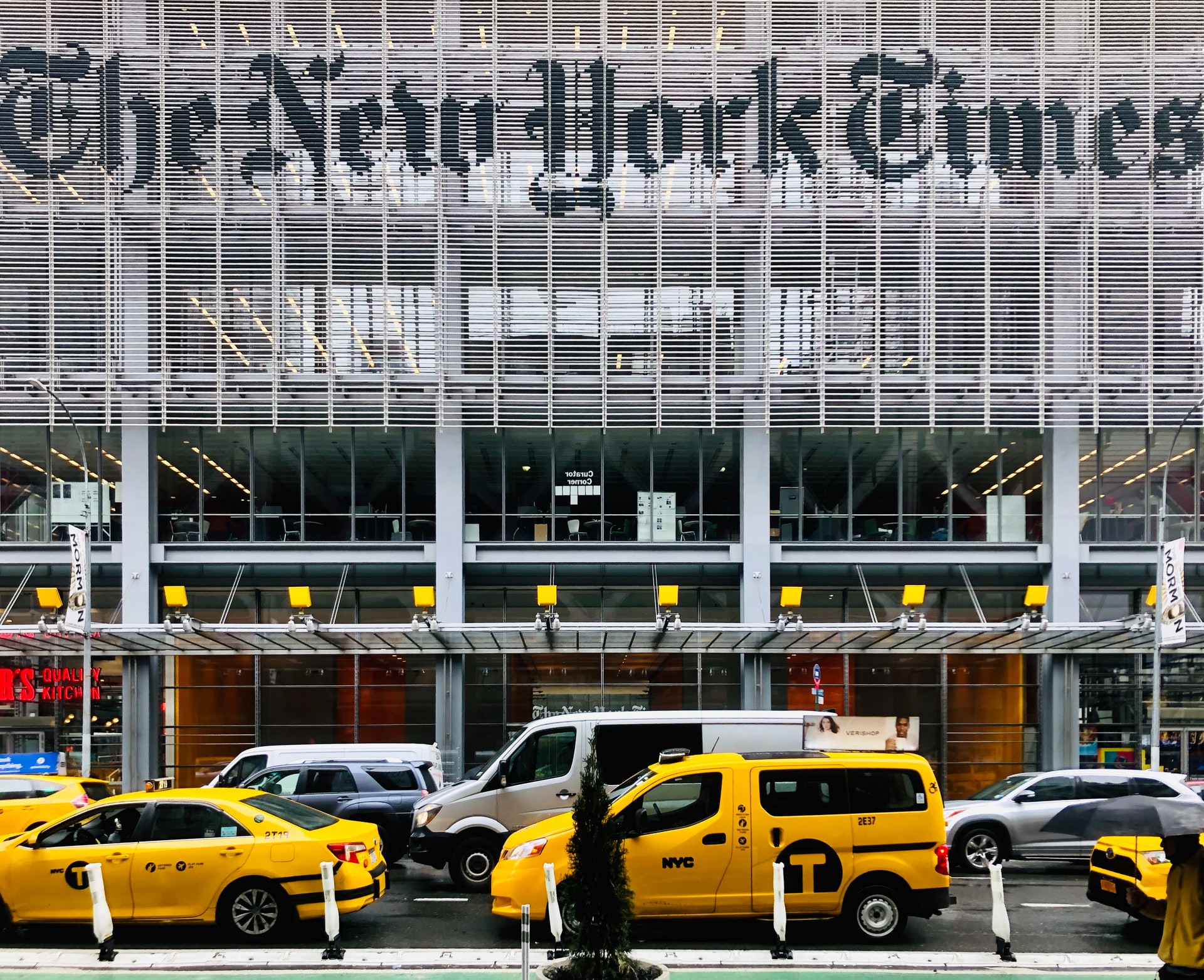 A view of facade of the New York Times Company, a grey metal and glass wall, with taxis and cars in front.