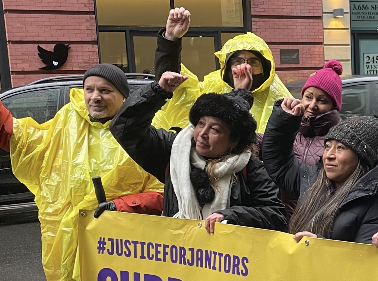 A group of workers in yellow rain jackets holing a banner in one hand that says hashtag justice for janitors and all raising their other fist high, standing in front of the NYC Twitter building and its logo on the wall in the background