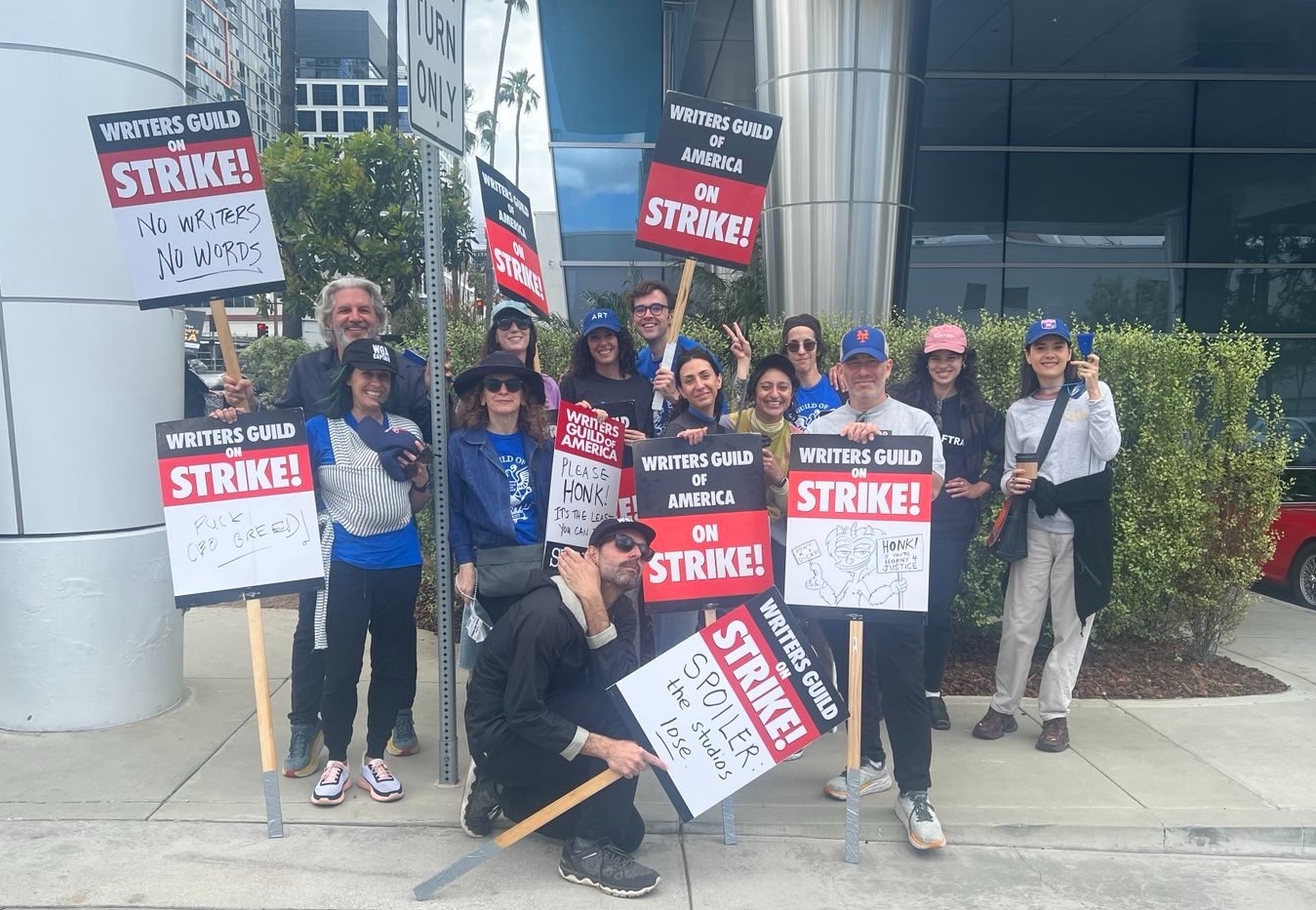 13 people standing and posing with Writers Guild placards in front of a grey corproate office building with palm trees in the background. Most have hats and glasses and are smiling despite the tough circumstances. Their placards say 'No Writers, No Words' and 'Spoiler: The Studios Lose' and more