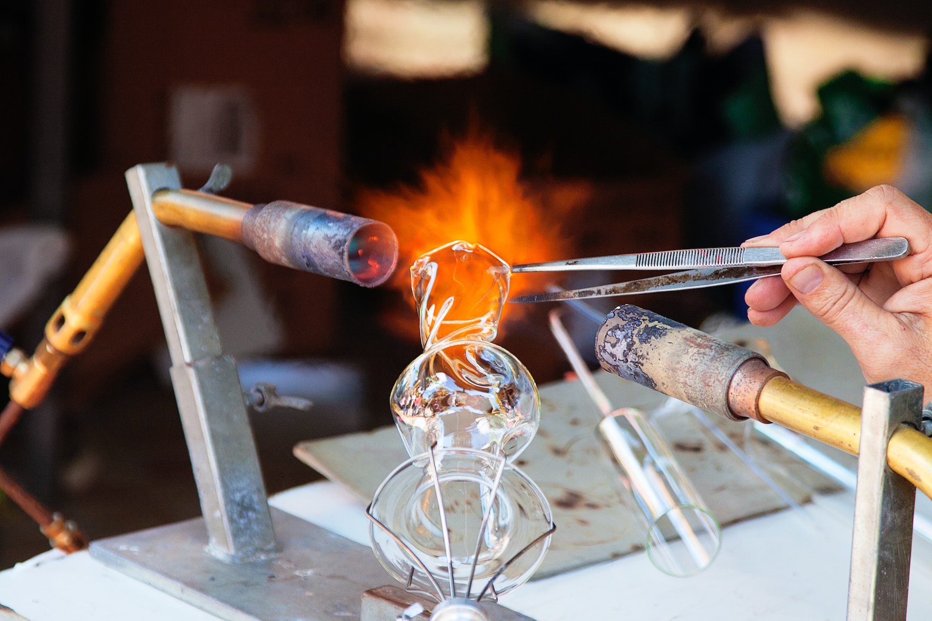 A piece of twisted glassware between two gas flames, the mouth on fire, and a hand on the right holding a pair of tweezers touching the top